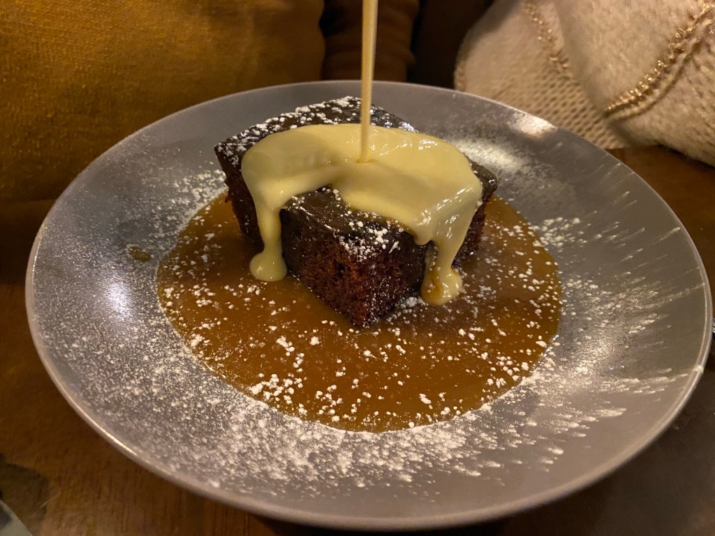 A plate with a piece of sticky toffee pudding, surrounded by toffee sauce and with custard being poured on 