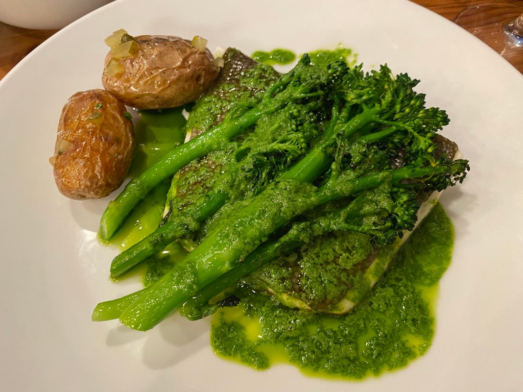 A piece of white fish underneath some long stem broccoli, drenched in a green parsley and butter sauce - with two potatoes