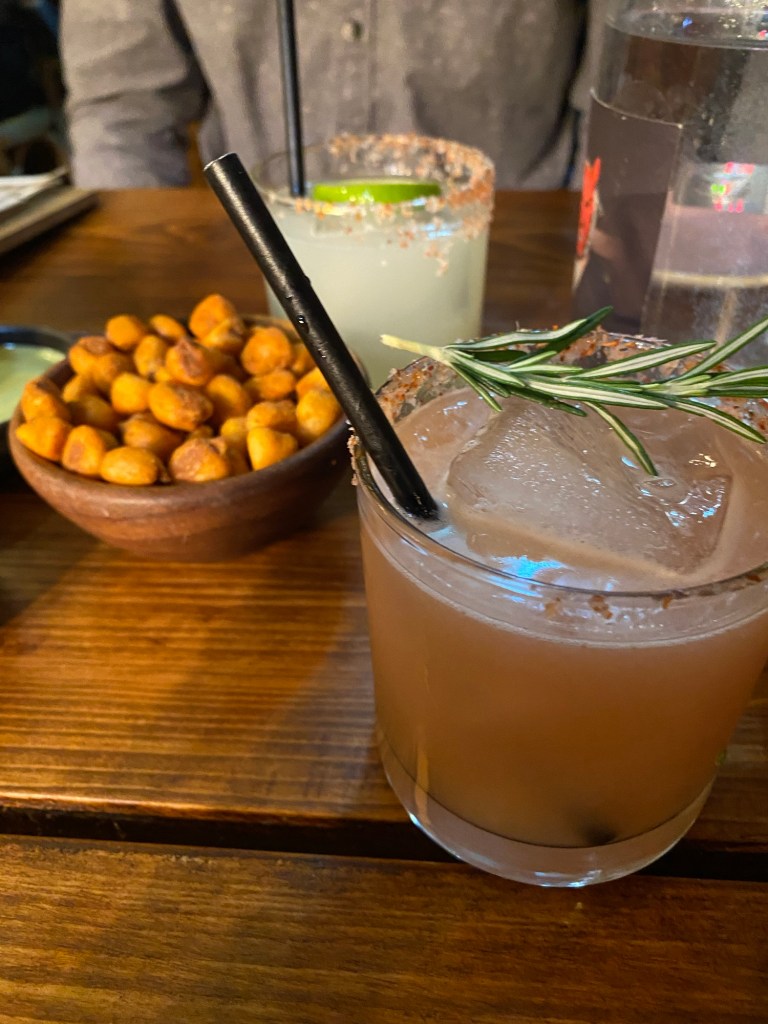 Two cocktails and a bowl of crunchy corn kernels on a wooden table