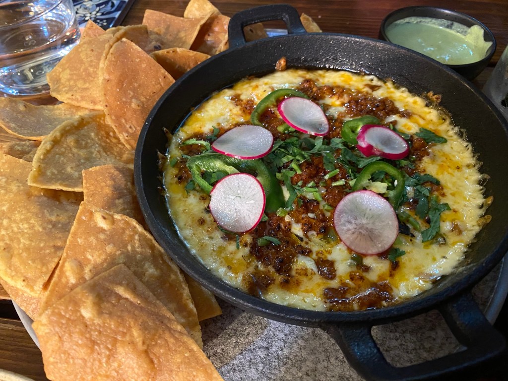 A skillet of melted cheese with radishes  and chorizo on top, and crunchy tacos on the side