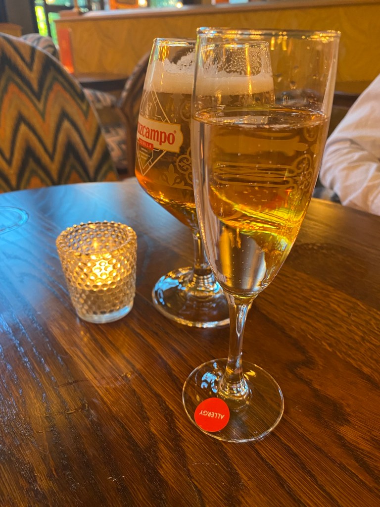 A glass of prosecco on a wooden table with an allergy sticker on it. Behind is a glass of beer and to the side a lit tealight in a holder