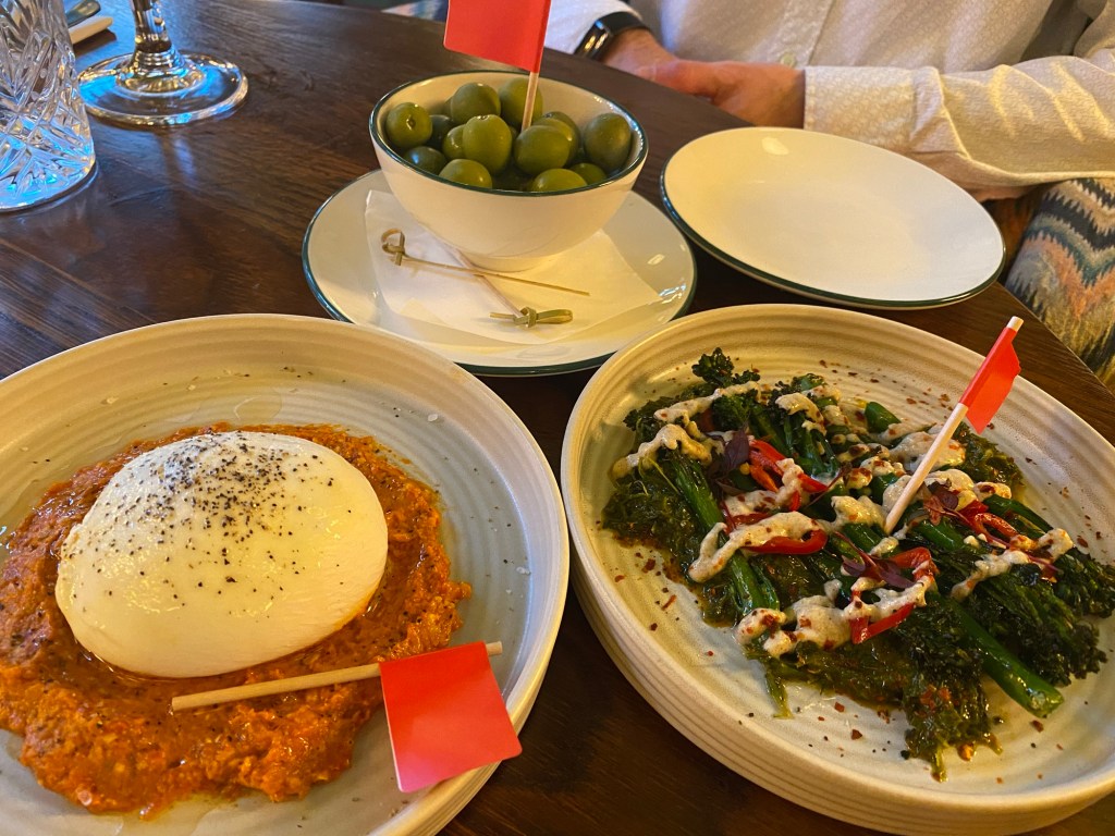 Three white serving bowls on a wooden table, containing olives, burrata cheese and tenderstem broccoli