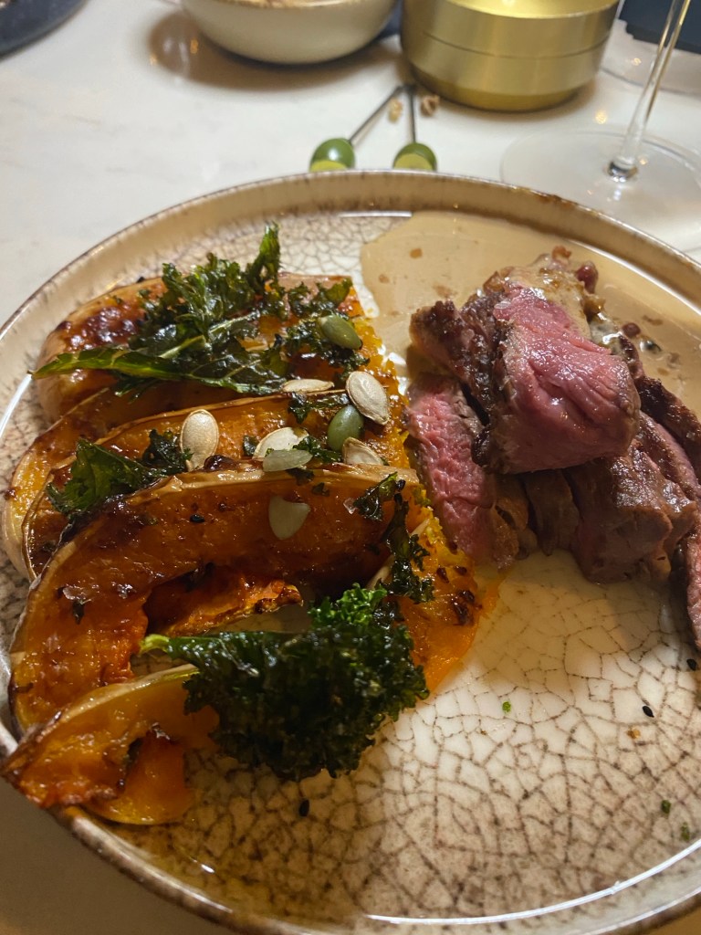 A round plate on a table containing, on the left, slices of baked squash topped with pumpkin seeds and kale, and on the right, slices of rare beef steak with peppercorn sauce