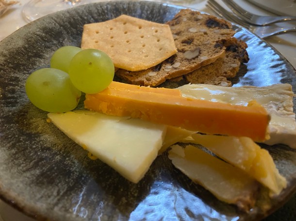 A plate with some slices of different coloured and textured cheeses, three green grapes and some crackers and toasts for cheese