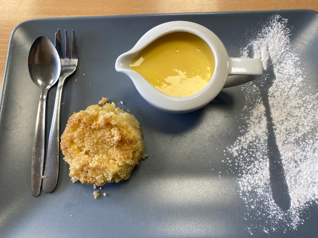 A rectangular, grey plate with a round apple crumble, a jug of custard, a spoon and fork. They have also made an outline of a fork as a decoration using icing sugar