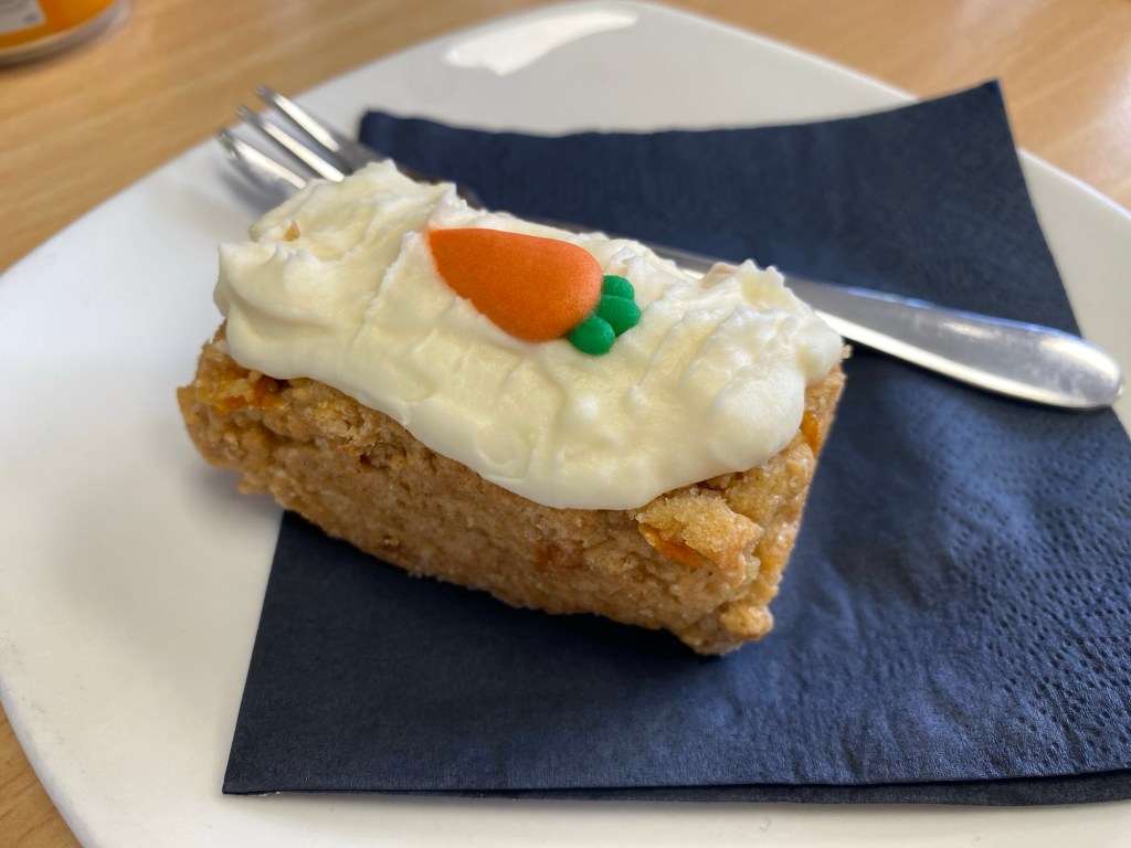 A piece of carrot cake with soft icing on the top and a miniature icing carrot, with a black paper napkin and a fork, on a white plate