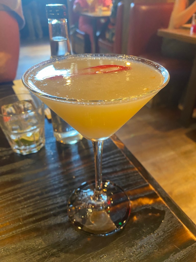 A martini glass with a spicy lychee margarita, a piece of red chilli floating in it and a salted rim, on a restaurant table