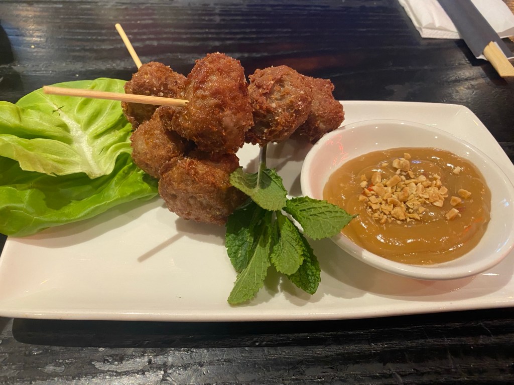 An oblong plate containing two skewers each with three pork and lemongrass meatballs at a jaunty angle, some pieces of mint and lettuce, and a small pot of peanut sauce