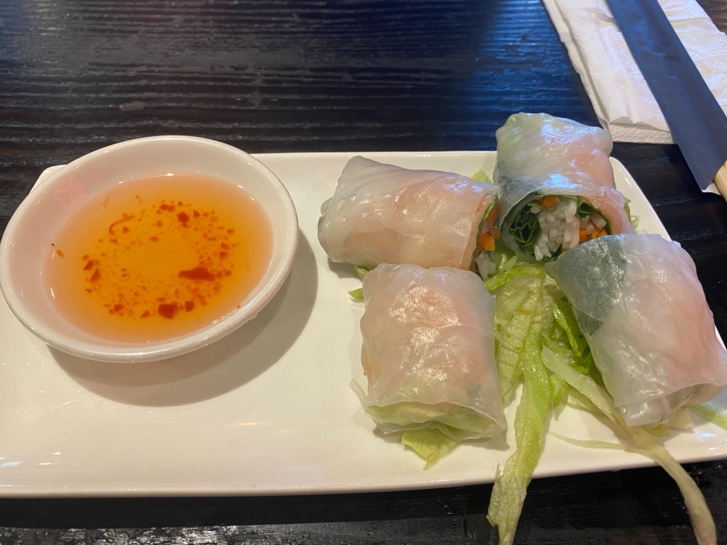 An oblong plate with two summer rolls cut in half on top of some shredded lettuce. To the left, a small pot of nuoc cham dipping sauce
