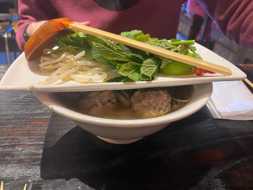 A large bowl of Pho soup, with a oblong plate on top containing a large wooden spoon and a big pile of herbs and beansprouts