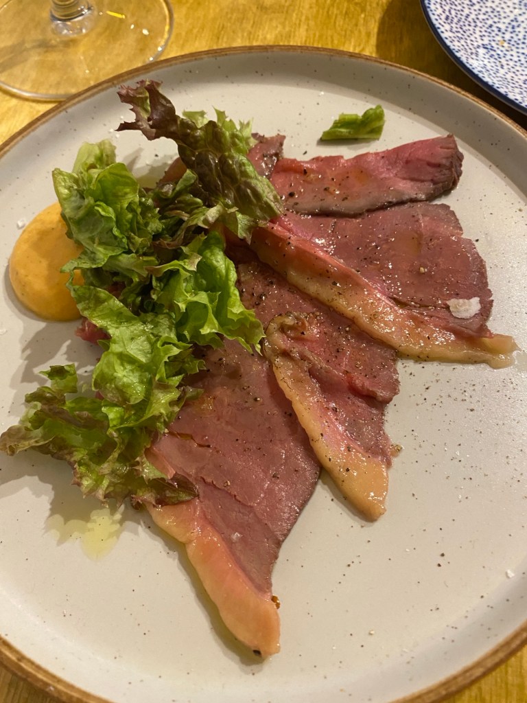 A plate containing very thin slices of barely cooked beef with some lettuce and a yellow goat cheese sauce 