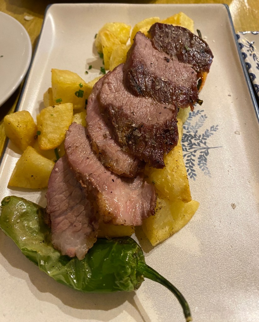 Slices of barbequed pork on top of some small roasted potatoes with a Padron pepper, on a cream coloured, rectangular plate
