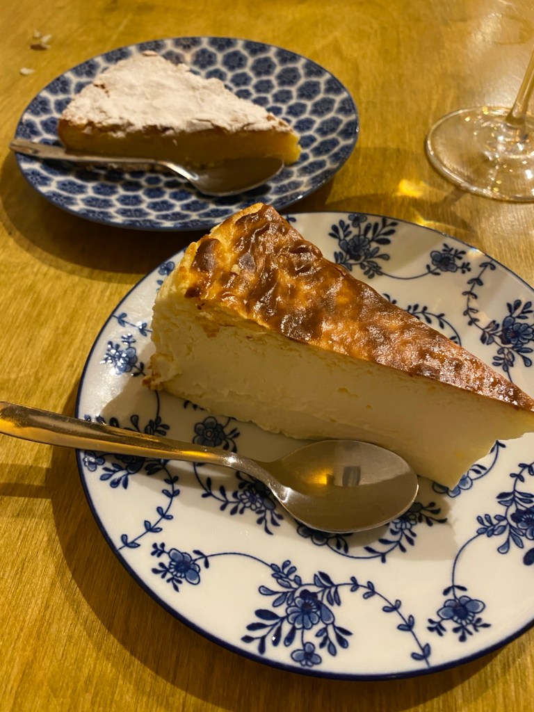 Two desserts on separate blue and white plates. In the foreground, a slice of burnt Basque cheesecake and behind, a slice of almond cake with icing sugar on the top