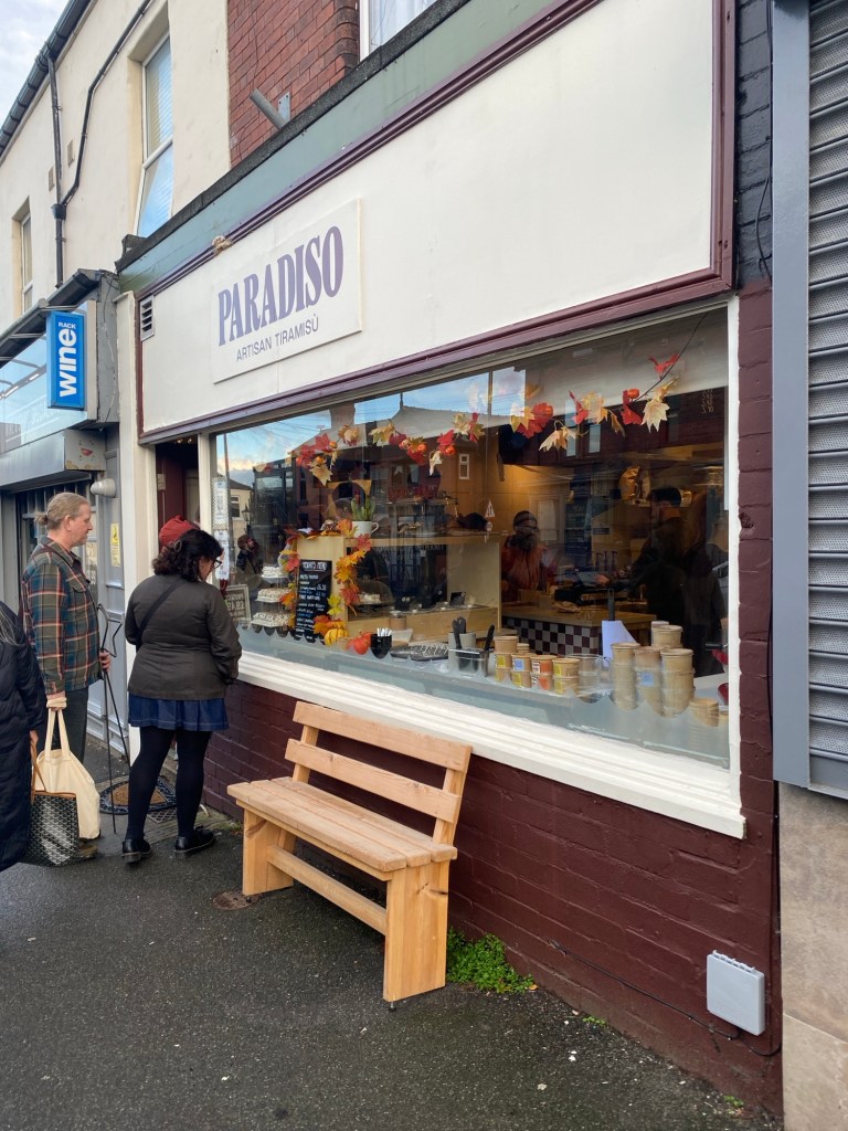 A view of the shop front of Paradiso Artisan Tiramisu on Sharrow Vale Road, Sheffield, with some pots of tiramisu visible in the window and a few people queueing outside