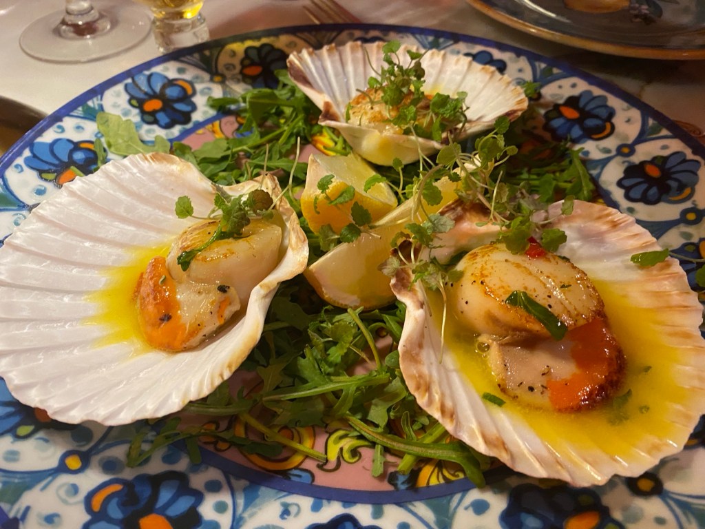 A plate with three scallop shells on a bed of green leaves, each containing a cooked scallop, topped with micro herbs and accompanied by a couple of lemon wedges