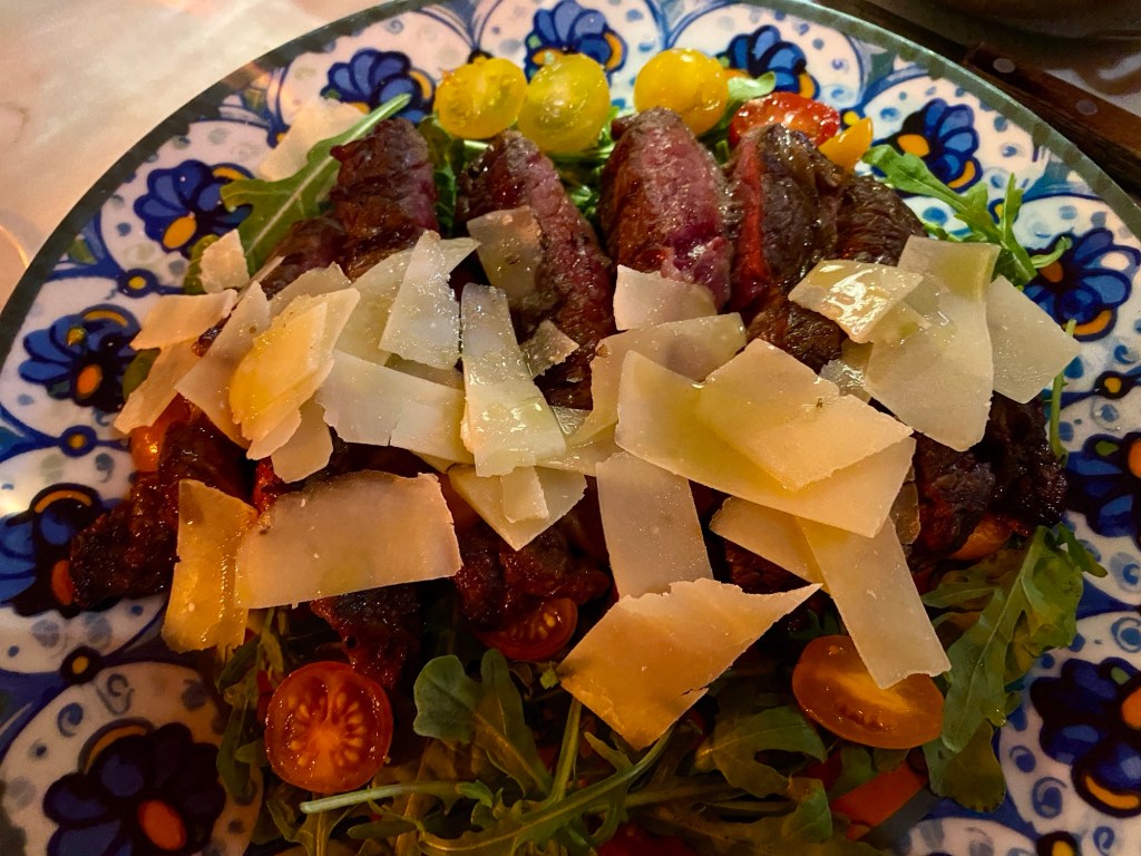 A plate with sliced ribeye beef and salad, hiding under a generous amount of large parmesan cheese shavings