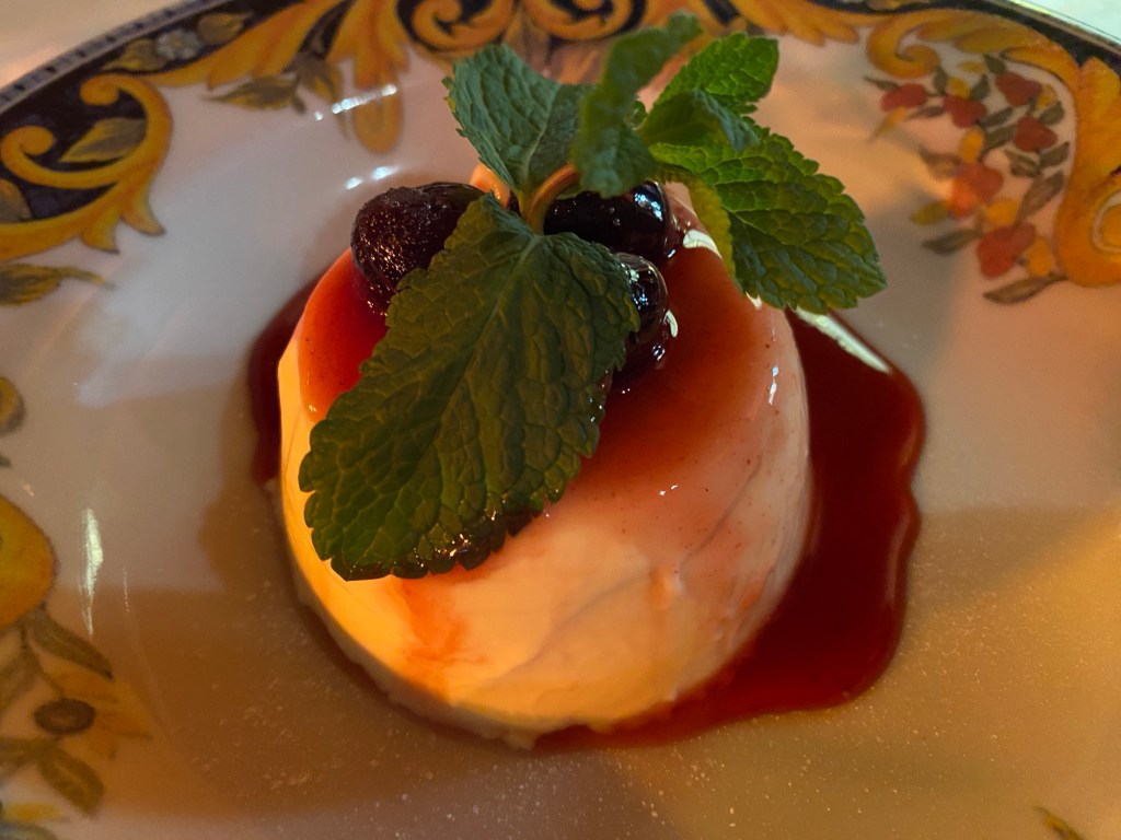 A decorative dish containing a pannacotta, topped with cherries and red, fruity sauce, and a sprig of mint leaves