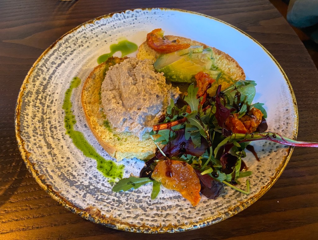 A pretty white and grey plate containing a piece of toast with crab pate on, slices of avocado, a side salad of greens and tomato, and a pretty, green drizzle