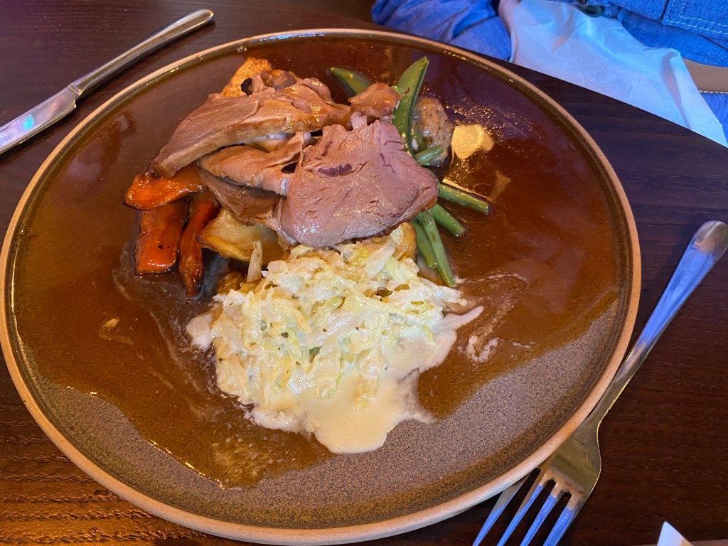 A brown plate with a classic Sunday roast with some of the trimmings (well, it is gluten free so you can't have all of them). We can see slices of roast lamb and a variety of vegetables hiding underneath it (carrots, parsnips, green beans) and what looks to be shredded celeriac in a white sauce 