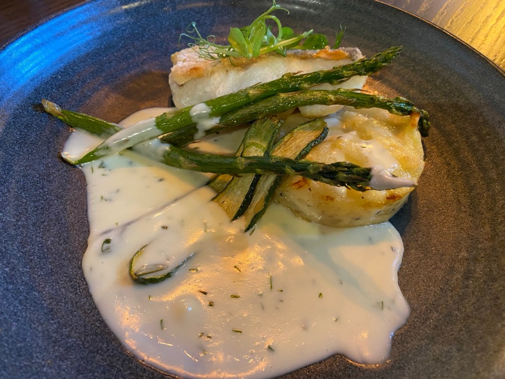 A grey plate containing a squat cylinder of potato gratin, upon which leans a piece of white fish with crispy skin, and some long pieces of asparagus and courgette strips. They all sit in a puddle of white sauce with herbs in.