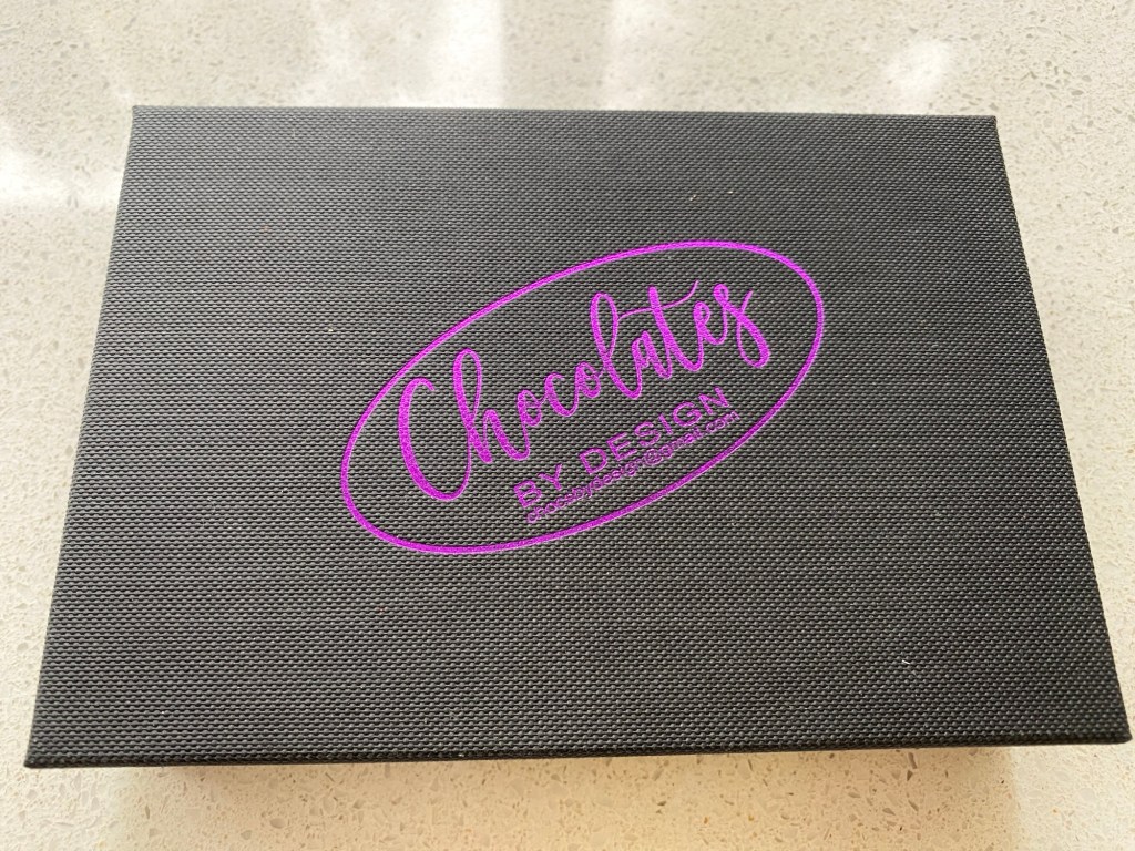 A rectangular, textured black box lid with purple writing saying Chocolates by Design 