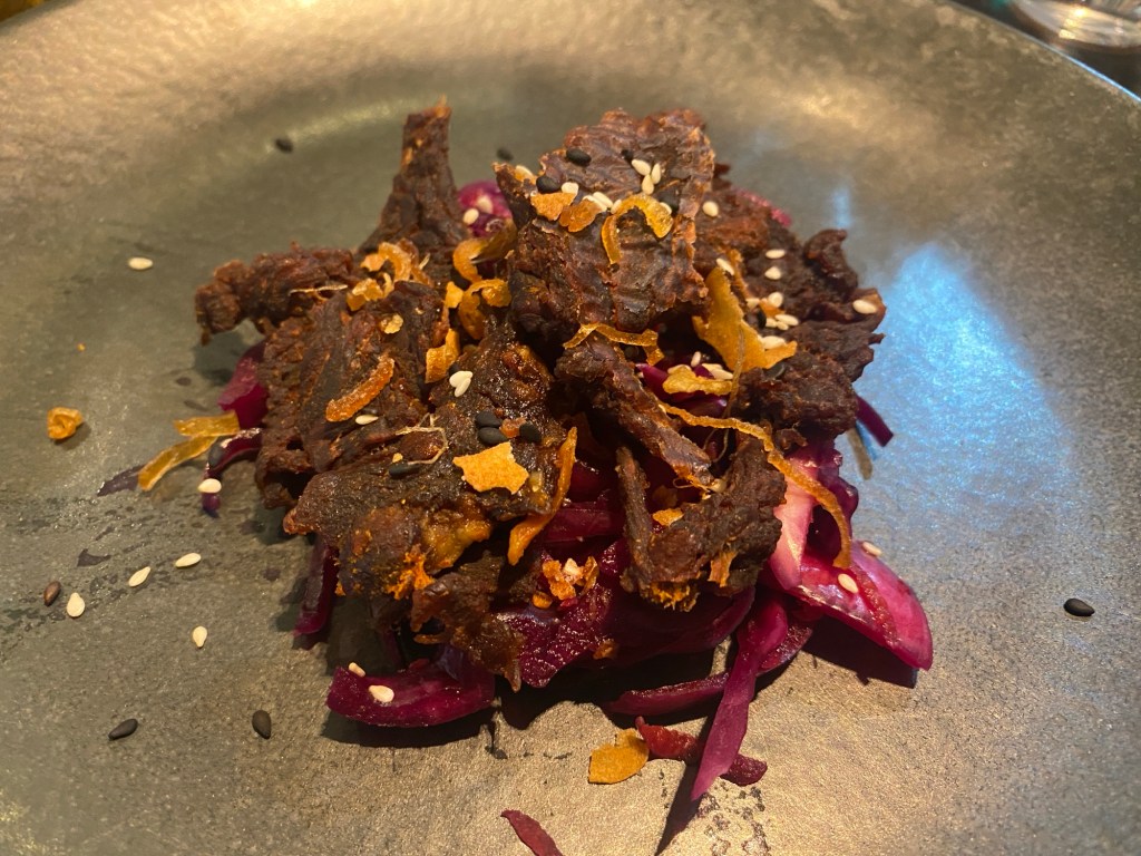 A pile of beef jerky sitting atop a purple slaw on a grey plate. Sprinkled with bits of crispy onion