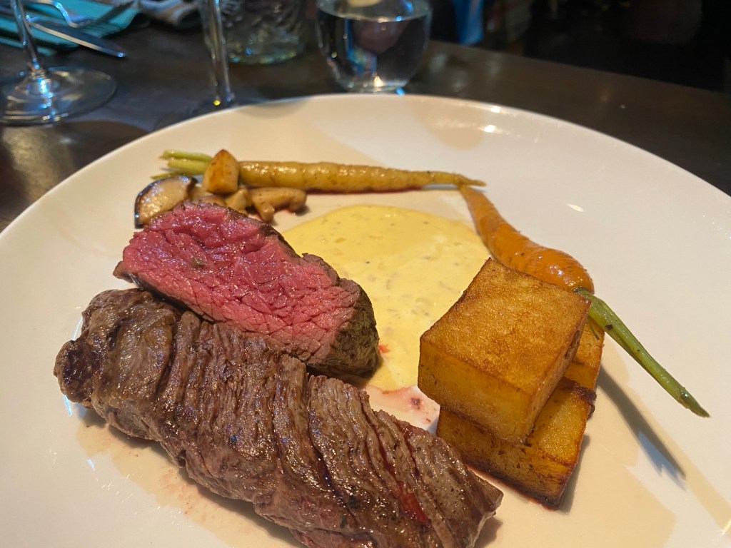 A white plate with two hunks of steak, one lovely and pink. Sitting alongside two large, crispy cubes of potato, some long thin carrots complete with greenery, a few mushrooms and a creamy Bernaise sauce