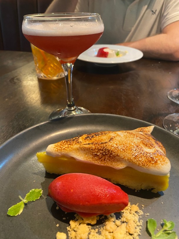 In the foreground, a luscious slice of lemon meringue pie is hiding behind a very beautiful quenelle of pink hibiscus sorbet. Behind the plate is a pink, foamy cocktail in a tall glass and in the background, someone else's pudding is out of focus.