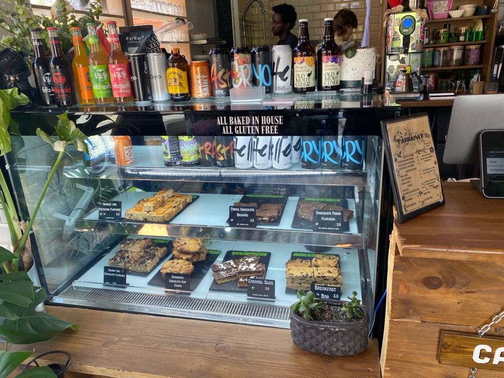A busy counter top in a cafe. On top of a wooden counter sits a tiered glass display containing a variety of tray bakes, all gluten free. On top of the display unit are a variety of bottled and canned drinks. A couple of plants are in the foreground.