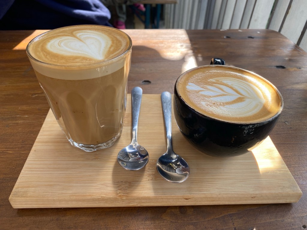 A small wooden tray on a darker wooden table. On it sits a glass of coffee on the left, with a frothy top and a heart shaped decoration in the foam. On the right a black cup of coffee with a swirly plant decoration in the foam two teaspoons sit in between the coffees. 