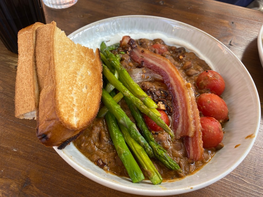 A white bowl on a wooden table containing lots of refried beans, and on top of that several spears of asparagus some streaky bacon and some small, cooked tomatoes.  
two pieces of thick, white bloomer toast sit perched on the left of the dish.