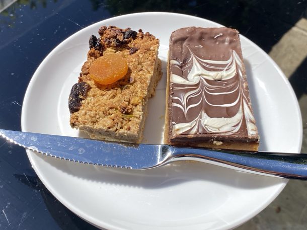 A white plate containing two slices of traybakes. On the left a fruity flapjack with raisins and, pumpkin seeds visible in the mix, and a dried apricot on top.
On the right is a slice of millionaire's shortbread, although we can only really see the top, which is milk chocolate with a swirly white chocolate pattern. In front, also on the plate, is a sharp, serrated knife  