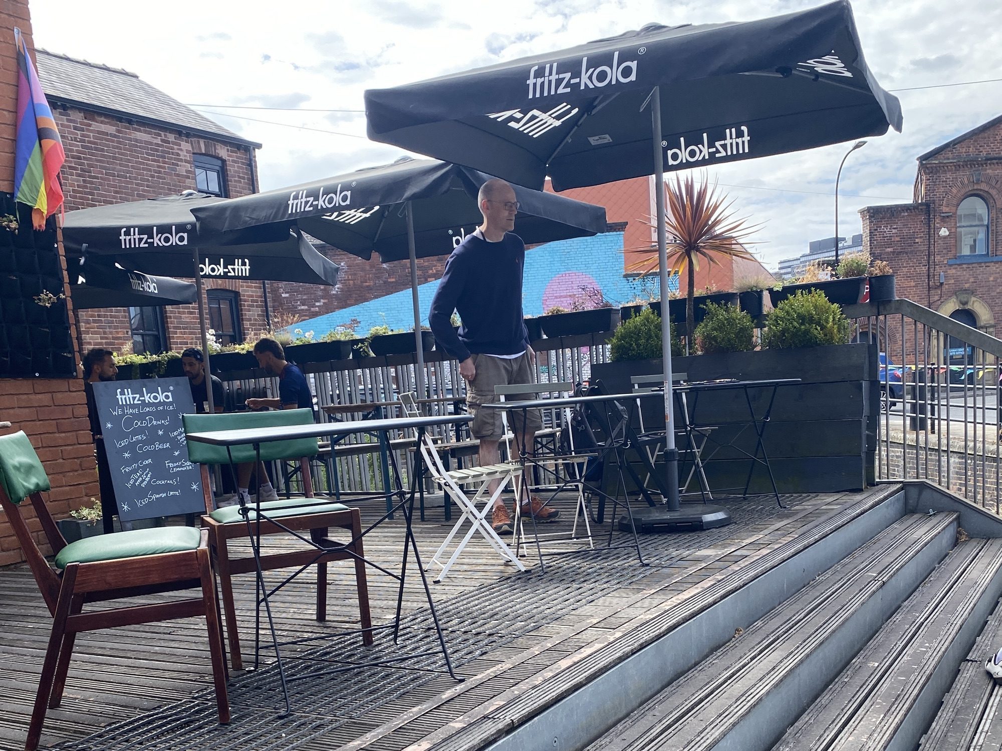 Taken at a slightly jaunty angle, this shows a raised decking, with a few steps at the right hand side. On the decking are chairs and tables under some big, black square umbrellas. A man is preparing to take a seat at one of the tables.